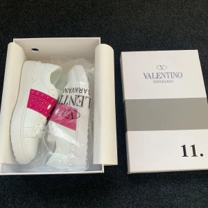 Valentino Garavani sneakers med rosa detalj - Snygga Valentino Garavani sneakers i vitt med en iögonfallande rosa detalj över sidan. Skorna kommer i originalförpackning och har en modern design som passar perfekt för en stilren look. köpte från plick och de är aldrig använda. vet ej om de är äkta.