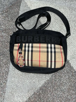 Burberry messenger bag - Snygg axelväska från Burberry med klassiskt beige rutigt mönster och svart tygdetalj där loggan är broderad. Väskan har justerbar axelrem och dragkedja med flätad detalj. Perfekt för dig som gillar stilrena accessoarer med lyxig känsla.