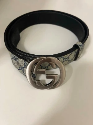 Gucci GG bälte med silverfärgat spänne - Snyggt bälte från Gucci med klassiskt GG-mönster i beige och blått tyg samt ett stort, silverfärgat GG-spänne. Perfekt accessoar för att lyfta din outfit med en lyxig touch och ikonisk design.