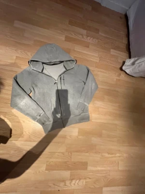 Grå zip hoodie från sail racing  - Snygg grå hoodie från sail racing med dragkedja framtill och huva. Tröjan har två fickor på magen och diskret logga på bröstet. Tillverkad i mjukt sweatshirtmaterial som är skönt att bära. Perfekt för en avslappnad stil.