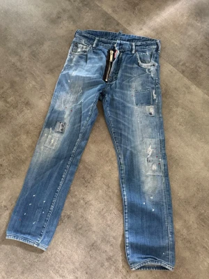 Blå patchade jeans med dragkedja - Säljer ett par blå jeans med patchade detaljer och slitningar, samt en synlig dragkedja framtill. Jeansen har raka ben och coola färgstänk för en trendig look. Perfekt för dig som gillar streetstyle och unika plagg.