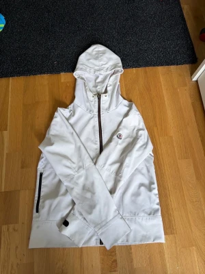 Vit vindjacka med huva från Moncler - Snygg vit vindjacka från Moncler med huva och hel dragkedja framtill. Jackan har svarta detaljer vid fickorna och en klassisk Moncler-logga på bröstet. Perfekt för blåsiga dagar och enkel att matcha med olika outfits.