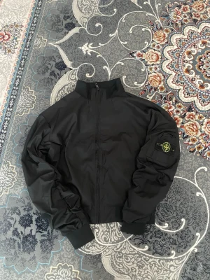 Svart jacka från Stone Island - Svart jacka från Stone Island med klassisk logotyp-patch på ärmen. Lätt, vindtät och perfekt för vår/sommar. Två ytterfickor, innerficka, samt armficka vid patchen. Sitter snyggt med den typiska Stone Island‑passformen. Materialet är slitstarkt och ger en modern streetwear-känsla.