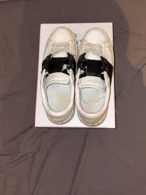 Valentino skor - Snygga vita sneakers från Valentino med en bred svart läderdetalj över mitten. Klassisk låg modell med vita skosnören och diskret logga på insidan. Tillverkade i läder för en exklusiv känsla och stilren look. En nackdel är att färgen har repats man märker inte så mycket när du har på dig de men annars lite smutsig sula men inget håll eller slipning på den knappt använd