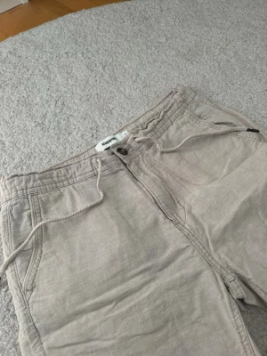 Beigea linneshorts med snörning från Kappahl - Snygga beige shorts från Kappahl med snörning i midjan och knappstängning. Tillverkade i ett mjukt bomullsmaterial och har två fickor fram. Perfekta för varma dagar och enkel att matcha med olika stilar.