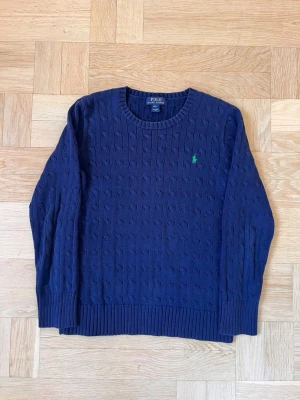 Mörkblå kabelstickad tröja Polo Ralph Lauren - Snygg mörkblå kabelstickad tröja från Polo Ralph Lauren med rund halsringning och grönt broderat logotyp på bröstet. Tröjan har ribbade muddar vid ärmslut och nederkant, perfekt för lager på lager och klassisk stil.