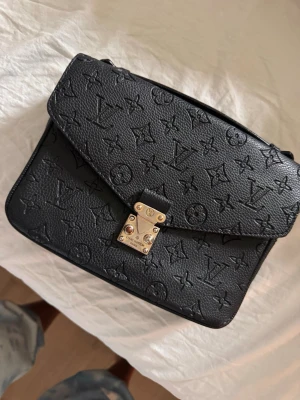 Svart handväska i läder - Snygg svart handväska  i präglat läder med klassiskt monogrammönster. Väskan har guldfärgat lås framtill och en dragkedja på baksidan. Perfekt storlek för det viktigaste och en stilren accessoar till din outfit. Obs se bilder och ingen axelrem medföljer. 