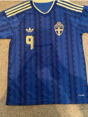 Blå Sverige fotbollströja Adidas - Säljer en blå Sverige fotbollströja från Adidas med gula detaljer, nummer 9 och namnet ISAK på ryggen. Tröjan har klassiska Adidas-ränder på axlarna, broderad landslagssköld och Climacool-material som andas. Perfekt för match eller träning.