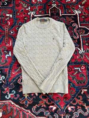 Beige kabelstickad tröja Morris - Snygg beige kabelstickad tröja från Morris med rund halsringning och diskret logga broderad på bröstet. Tröjan har långa ärmar och ribbade muddar vid ärmslut och nederkant. Perfekt för lager på lager och klassisk stil. Tröjan är i storlek S. Tänk på att Morris storlekar ibland kan vara pytte lite mindre än andra märken i samma storlek. PRISET KAN DISKUTERAS!
