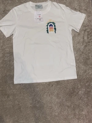 Vit t-shirt från Casablanca med tryck - Vit t-shirt från Casablanca med färgglatt tryck av ett fönster och byggnad på ryggen samt mindre tryck på bröstet. Klassisk passform och rund halsringning. Perfekt för dig som gillar unika och konstnärliga motiv.