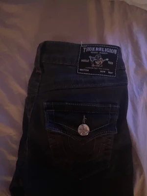 True Religion grå bootcut jeans - Grå  jeans från True Religion med bootcut-modell och snygga detaljer på bakfickan, inklusive en silverfärgad knapp och kontrastsömmar. Klassisk femficksdesign och tydlig logopatch bak. Perfekta för en trendig och avslappnad look.