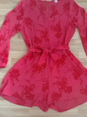 Som ny. Röd blommig jumpsuit med knytband - Hade på mig bara en gång. Snygg röd jumpsuit med blommigt mönster och långa ärmar. Modellen har ett matchande knytband i midjan och dragkedja bak. Materialet är lätt och flowy, perfekt för en trendig look. Passar dig som vill sticka ut med färg och mönster. Storlek Xl, men förminskad som M, som HM storlekar brukar vara. Bröst 102cm, midjan 85cm, längd 82cm.