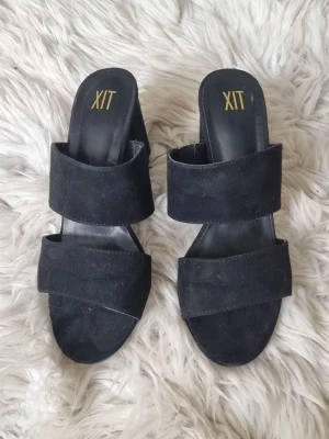 Svarta klackade sandaler från XIT - Stilrena svarta sandaler från XIT med två breda remmar över foten och öppen tå. Skorna har en hög, fyrkantig klack och ovandel i mockaimitation. Perfekta för dig som vill ha en trendig och bekväm look.
