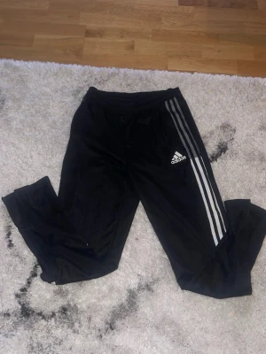 Svarta Adidas träningsbyxor - Säljer ett par svarta träningsbyxor från Adidas med de klassiska tre vita ränderna längs benen och Adidas-logga på låret. Byxorna har resår i midjan och är tillverkade i ett mjukt, lätt material som passar perfekt till träning eller chill. Står inte vilken storlek