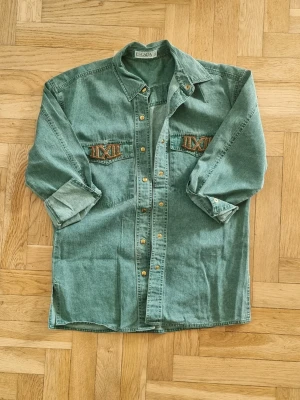Grön jeansjacka från Escada - Vintage grön jeansjacka från Escada med klassisk krage och tryckknappar framtill. Två bröstfickor med unika broderade detaljer i brunt och guld. Avslappnad passform och långa ärmar som kan vikas upp för en cool look.