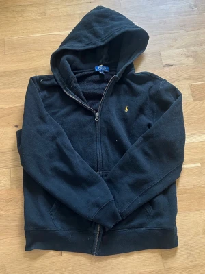 Svart hoodie från Polo Ralph Lauren - Svart hoodie med dragkedja från Polo Ralph Lauren. Klassisk design med huva, fickor framtill och den ikoniska gula loggan broderad på bröstet. Mjuk insida och skön passform, perfekt för en avslappnad stil.