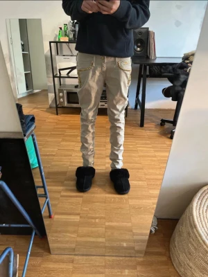 Beiga cargobyxor med kedjedetaljer - Säljer ett par beiga robin’s jean med slim fit och snygga nitar vid fickorna. Byxorna har flera fickor på sidorna.