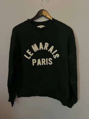 Svart sweatshirt Le Marais Paris - Cool svart sweatshirt med vit text 'LE MARAIS PARIS' på bröstet. Klassisk rund halsringning och långa ärmar. Perfekt för en avslappnad streetstyle-look.
