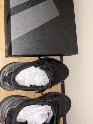 Svarta Adidas sneakers med box - Snygga svarta Adidas sneakers med klassiska tre ränder på sidan. Skorna har en låg profil och är tillverkade i textil och syntetmaterial. Perfekta för en sportig och stilren look. Levereras med originalkartong.