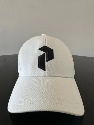 PeakPerformance Vit keps storleks/M, flex - Snygg vit keps med böjd skärm och en stilren svart logotyp framtill. Kepsen har klassisk sexpanelsdesign och är tillverkad i ett slitstarkt bomullstyg. Perfekt accessoar för en clean och modern look.
