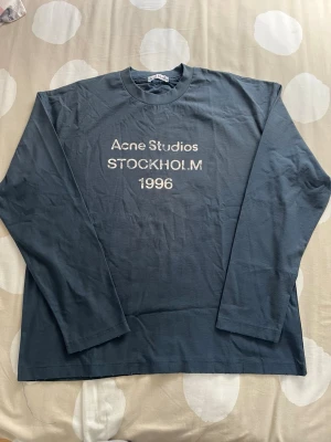 Marinblå långärmad Acne Studios  - Snygg blå långärmad  från Acne Studios med tryck 'Acne Studios STOCKHOLM 1996' på bröstet. Tillverkad i mjuk bomull och har en klassisk rund halsringning. Perfekt för en clean och avslappnad stil.