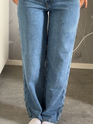 Blå raka jeans med hög midja - Säljer ett par klassiska blå jeans med rak passform och hög midja. Jeansen har fem fickor, dragkedjegylf och är tillverkade i denim. Perfekta för en avslappnad och trendig look.