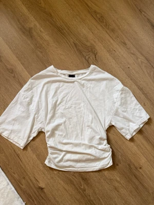 Vit t-shirt från Gina Tricot - En basic vit t-shirt från Gina Tricot med korta ärmar och rund hals. T-shirten är i mjuk bomull och har en lätt loose passform med en liten twist i midjan för en cool look.