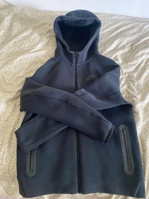 Nike tech - Snygg svart hoodie från Nike med hel dragkedja och två fickor med dragkedja. Hoodien har en klassisk huva och diskret Nike-logga på bröstet. Perfekt för en sportig och avslappnad stil. Materialet är mjukt och bekvämt.