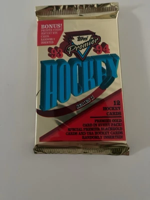Topps Premier Hockeykort 93/94 oöppnat - Oöppnat paket med 12 Topps Premier hockeykort från säsongen 1993/94. Förpackningen är guldig med blå och röda detaljer. Innehåller chans på premiumpremiumkort och specialkort. Perfekt för samlare av sportkort och hockeymemorabilia.