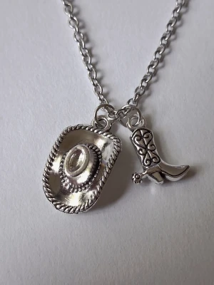 Silverfärgat halsband med cowboytema - Unikt halsband i silverfärg med två berlocker: en cowboyhatt och en detaljerad cowboyboot. Kedjan är tunn och smidig, perfekt för dig som gillar westernstil och vill sticka ut med coola accessoarer. Materialet är rostfritt metall med fina detaljer på berlockerna.