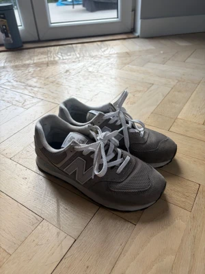 Grå New Balance sneakers - Snygga grå sneakers från New Balance med klassisk N-logga på sidan. Skorna har vita snören, meshpaneler för extra ventilation och detaljer i mocka. Perfekta för en avslappnad streetstyle-look och riktigt bekväma att gå i.