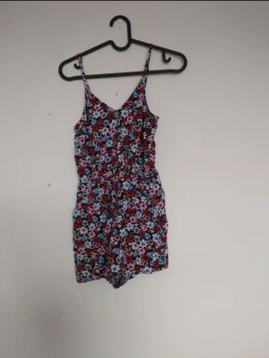 Blommig jumpsuit med smala axelband - Snygg blommig jumpsuit med smala axelband och V-ringning. Mönstret består av röda, blå och vita blommor på svart botten. Resår i midjan för en skön passform. Perfekt för varma dagar och enkel att styla med sneakers eller sandaler.