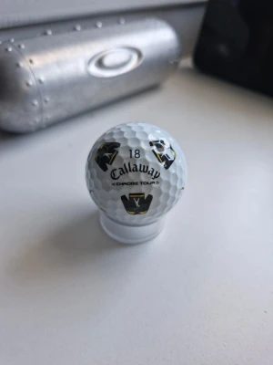 Callaway Happy gilmore +kartong - Säljer en vit Callaway Chrome Tour golfboll med svarta och gula V-mönster och siffran 18 tryckt på. Bollen har klassisk dimple-yta och är perfekt för dig som gillar golf och vill ha en boll med snygg design. Är från happy gilmore filmen