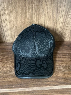 Svart keps från  med GG-mönster - Snygg svart keps  med det klassiska GG-mönstret över hela kepsen. Kepsen har böjd skärm och justerbar rem baktill. Tillverkad i ett slitstarkt tygmaterial med läderdetaljer längs skärmen. Perfekt för en trendig streetstyle-look.