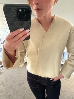 Beige omlottkofta från Gina Tricot - Stilren beige omlottkofta från Gina Tricot med v-ringad hals och breda ärmslut. Koftan har en rak, något kortare passform och dekorativa sömmar längs kanterna. Perfekt att matcha med jeans eller kjol för en trendig look.