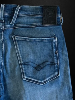  Replay Anbass - Säljer ett par blå slim fit jeans från Replay med klassisk femficksdesign och snygga slitningar. Jeansen har en mörk tvätt och diskreta kontrastsömmar. Bakfickan har Replay-logga och unik söm. Perfekta för en trendig look.