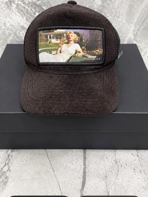 Svart keps från Stacchore med mesh - Säljer en svart keps från Stacchore med frottéliknande tyg framtill och mesh baktill. Framtill finns en stor patch med ett retroinspirerat motiv av en kvinna i solstol. Sidan har en metallogo. Justerbar baktill för perfekt passform.