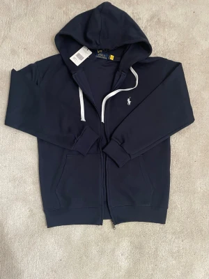 Marinblå kofta från Polo Ralph Lauren - Snygg marinblå hoodie från Polo Ralph Lauren med dragkedja, vita snören och klassisk logga broderad på bröstet. Tillverkad i mjukt bomullsmaterial och har två fickor framtill samt ribbade muddar. Perfekt för en avslappnad och stilren look.