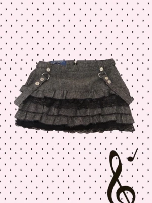 Grå volangkjol från Jennyfer - Kort grå kjol från Jennyfer med volanger och dekorativa knappar framtill. Kjolen har ett diskret mönster, svarta spetsdetaljer och metallringar som ger en cool touch. Perfekt för dig som gillar en edgy och unik stil.
