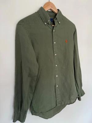 Olivgrön skjorta från Ralph Lauren - Klassisk olivgrön skjorta från Ralph Lauren med orange broderad logga på bröstet. Storleken är XS i amerikansk storlek, motsvarande S. Köpt i USA. Använd >10 gånger, passade ej.