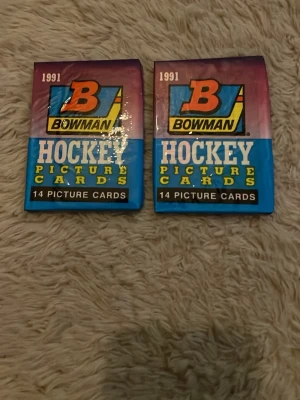 Bowman Hockeykort 1991 oöppnade - Två oöppnade paket Bowman Hockey Picture Cards från 1991. Varje paket innehåller 14 samlarkort. Förpackningarna är i färgerna blå, lila och orange med tydlig logga och text. Perfekt för dig som gillar hockey eller samlar på sportkort.
