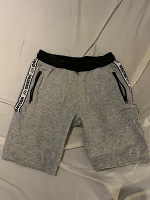 Grå shorts med svarta detaljer - Snygga grå shorts från WAIKND med svarta dragkedjor på fickorna och svart resår i midjan. Sidorna har vita band med svart WAIKND-tryck. Perfekta för en avslappnad stil och passar till många tillfällen. Storlek 158.