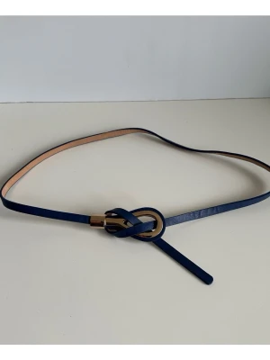 Mörkblått bälte med gulddetalj - storlek S - Snyggt bälte med en unik knutdetalj och guldfärgat spänne, storlek S. Bältet är 0,5cm smalt, 107cm långt och har en slät yta. Det guldfärgade spännet är designat för att enkelt fästas genom en ögla, vilket skapar en stilren och justerbar passform. Mycket gott skick, som ny!