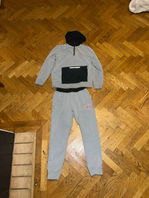 Grått loungewear-set från Under Armour - Mysigt grått loungewear-set från Under Armour med svart huva och magficka. Tröjan har dragkedja vid halsen och logga på bröstet, byxorna har resår i midjan och logga på låret. Perfekt för chill dagar hemma.