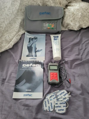 Cefar Primo TENS-apparat med tillbehör - Elektronisk TENS-apparat från Cefar med röd och svart kontrollenhet, fyra självhäftande elektroder, gel, manualer och grå förvaringsväska. Perfekt för smärtlindring och muskelstimulering. Allt samlat i ett praktiskt kit.