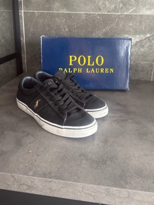 Svarta Polo Ralph Lauren sneakers - Svarta sneakers från Polo Ralph Lauren med klassisk låg profil, vita sulor och svart textilmaterial. Ikoniska Polo-loggan i guld på sidan och Polo-text på plösen. Snygga och stilrena skor som passar till det mesta.