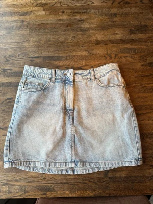 Ljusblå jeanskjol i denim - Trendig ljusblå jeanskjol i kort modell med klassiska fickor fram och bälteshällor. Kjolent har knapp och dragkedja framtill och är tillverkad i ett slitstarkt denimtyg. Perfekt för en avslappnad och cool stil.