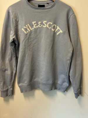 Ljusblå sweatshirt från Lyle & Scott - Ljusblå sweatshirt med rund hals och broderad Lyle & Scott-logga i vitt framtill. Tröjan har ribbade muddar vid ärmslut och nederkant, samt en avslappnad passform. Perfekt för en chill och stilren look.
