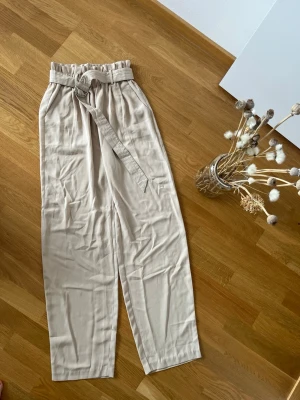 Beige byxor med papperspåse-midja - Stilrena beige byxor med papperspåse-midja och tillhörande knytskärp. Byxorna har vida ben och sidofickor, vilket ger en avslappnad och trendig look. Perfekta för en chill och snygg outfit under varma dagar.