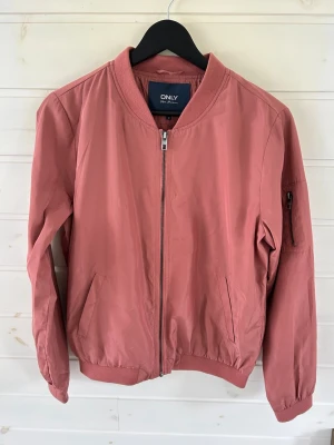 Rosa bomberjacka från ONLY - Snygg rosa bomberjacka från ONLY med dragkedja framtill, ribbade muddar och fickor på sidorna. Jackan har även en ärmficka med dragkedja och klassisk bomberkrage. Perfekt för dig som vill ha en stilren och trendig look.